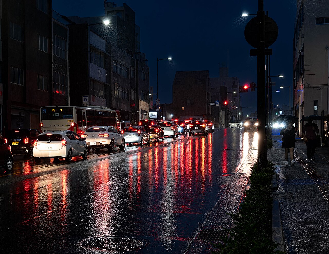 A wet Kanazawa street at dusk, with car lights reflected on the road and people walking under umbrellas